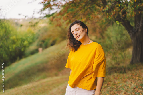 A woman in a bright yellow top walks through a park, smiling happily as she enjoys the beauty of nature and the warmth of the sun in autumn.