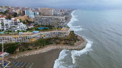 Drone flight along the coastline of Torremolinos overlooking the Bajondillo and Carihuela beaches, residential sector. Stormy weather, waves at sea, grey sky.