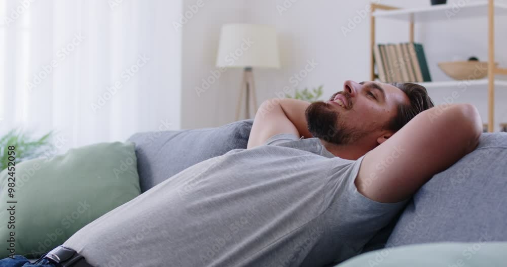 Smiling bearded Caucasian man relaxing on couch at home, stretching with his hands behind his head. He looking up with satisfied expression, enjoying peaceful break from work in his cozy living room. Smiling bearded Caucasian man relaxing on couch at home, stretching with his hands behind his head. He looking up with satisfied expression, enjoying peaceful break from work in his cozy living room.