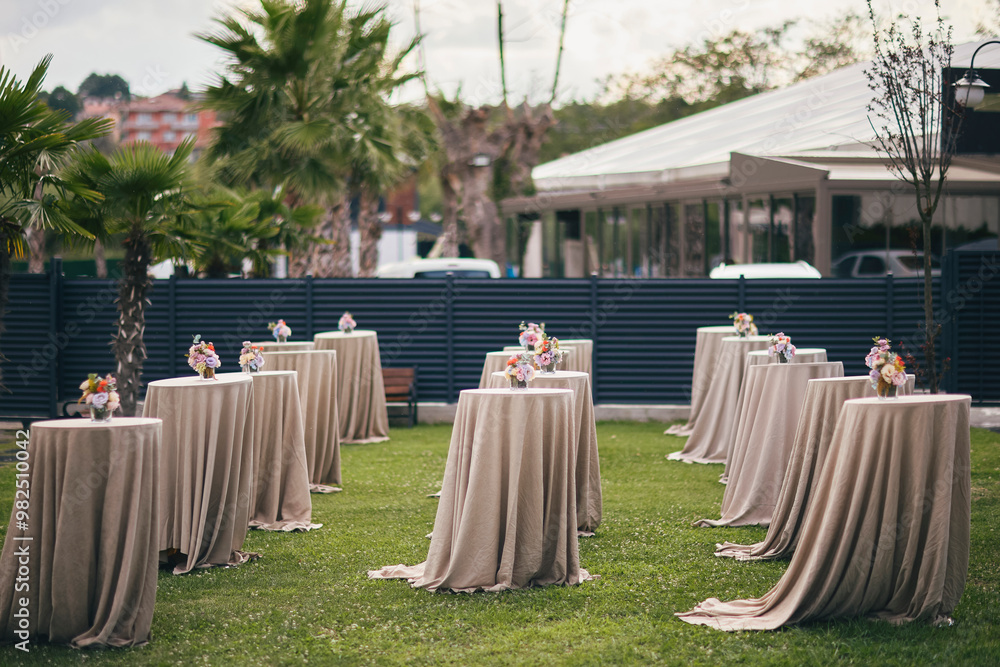 A stylish outdoor event space with round tables, lush greenery, and colorful floral arrangements