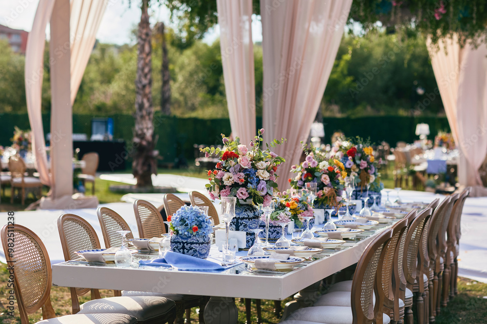 A long, elegant wedding table decorated with colorful floral arrangements, white linens, and gold flatware, set against a backdrop of lush greenery