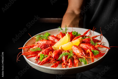 A plate of red boiled crayfish garnished with lemon wedges and herbs, held by a person against a dark background. Perfect for culinary, seafood, and gourmet food themes. photo