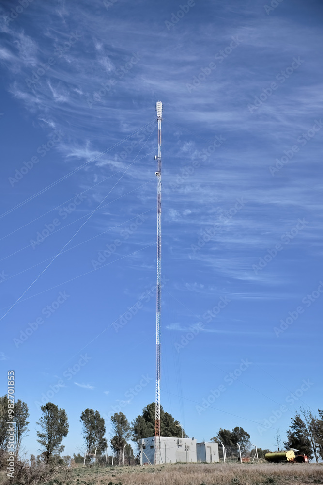 Obraz premium Tall TV communication tower with antennas against a blue sky and green field