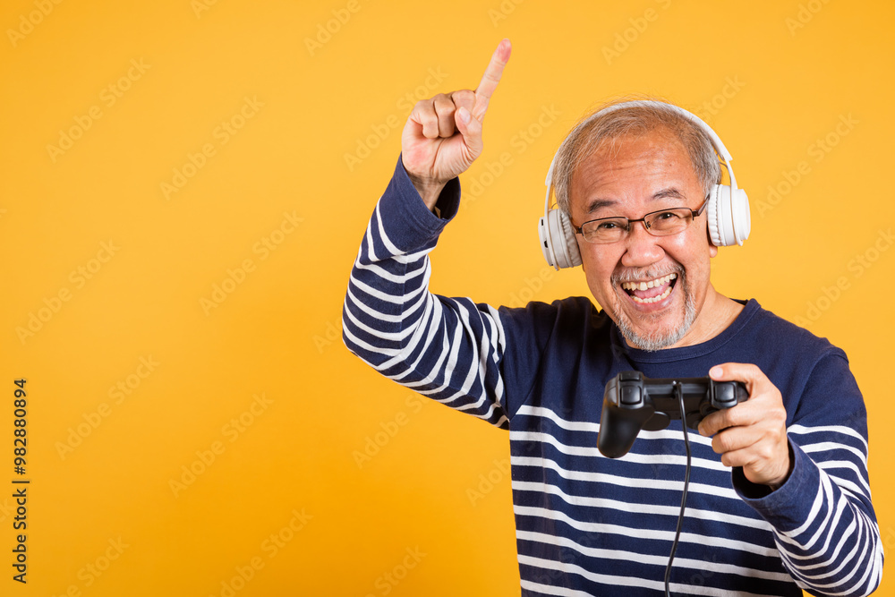 Portrait Asian smiling old man with headphones holding game controller studio shot isolated yellow background, senior man pensioner feeling winning playing a video game with joystick console Portrait Asian smiling old man with headphones holding game controller studio shot isolated yellow background, senior man pensioner feeling winning playing a video game with joystick console
