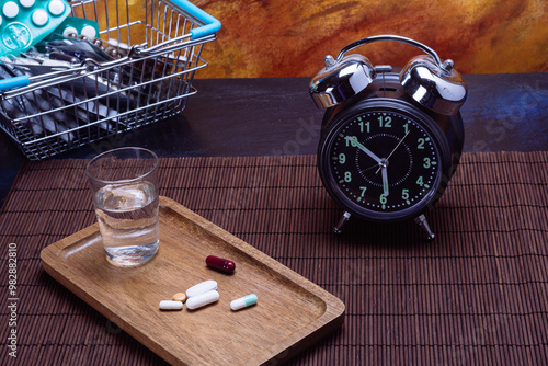 Variety of pills on tray with water, alarm clock, and shopping basket photo