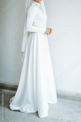 A woman, elegantly adorned in a stunning white dress and a delicate veil, stands gracefully in front of a pristine white wall, creating a beautiful and serene atmosphere around her