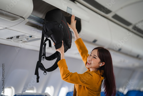 Woman Storing Luggage in Airplane Overhead Compartment: A young woman in a mustard yellow blazer smiles as she secures her black backpack in the overhead compartment of an airplane. The image captures