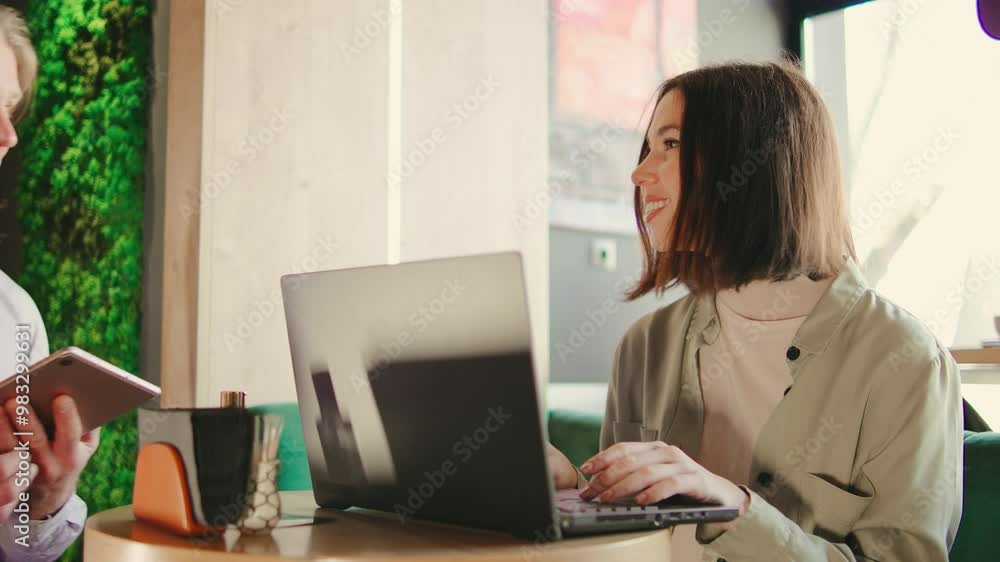Brunette woman works on her laptop at a coffee shop table while a waiter approaches with a tablet to take her order Brunette woman works on her laptop at a coffee shop table while a waiter approaches with a tablet to take her order
