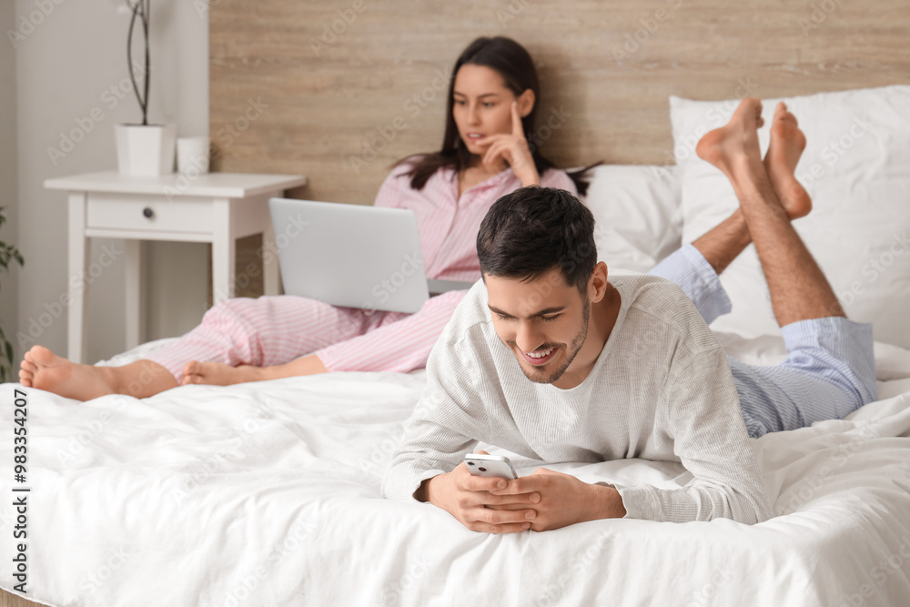 Young guy with mobile phone and his girlfriend with laptop lying on bed Young guy with mobile phone and his girlfriend with laptop lying on bed
