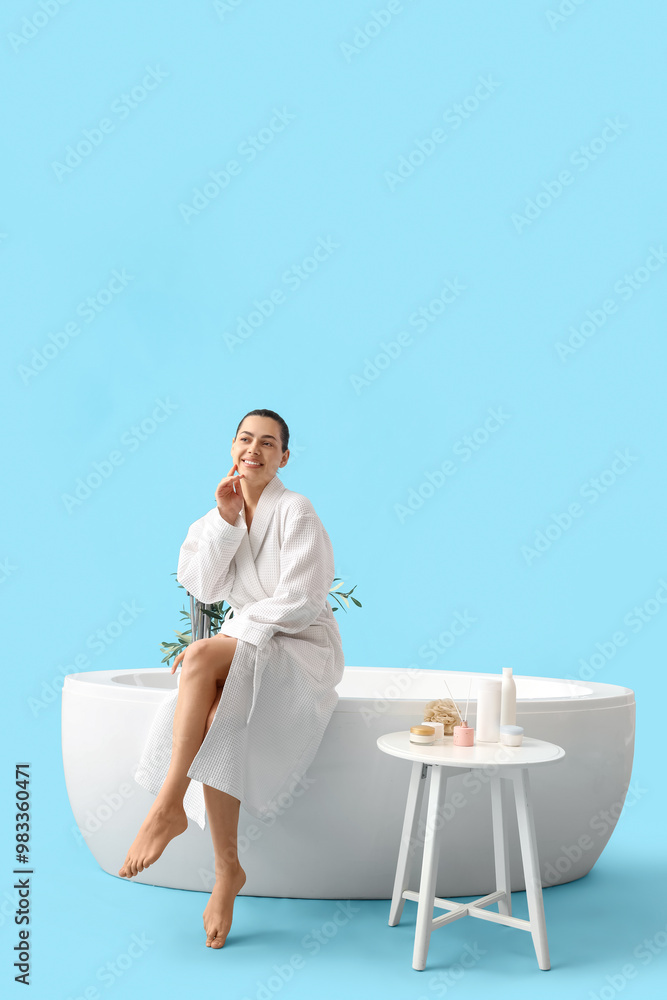 Young woman in bathrobe sitting on bathtub against blue background Young woman in bathrobe sitting on bathtub against blue background