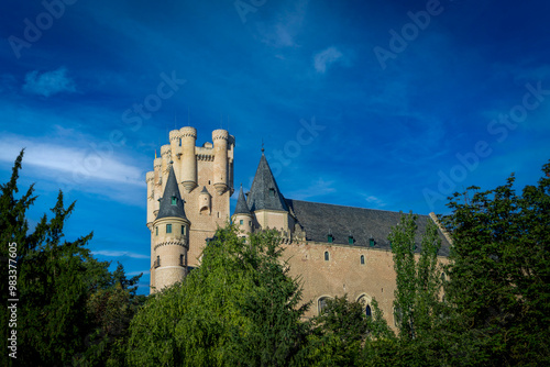 View of the north face of the Alcazar of Segovia, Castilla y Leon, Spain, from the Paseo del Eresma with its thick grove of trees and intense blue sky