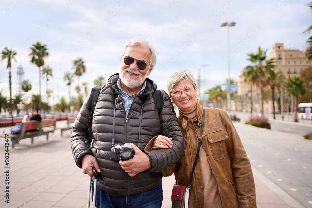 Portrait of smiling Caucasian senior tourist couple standing posing looking at camera in European street with their luggage. Retired people enjoying leisure time in winter holidays on sunny day Portrait of smiling Caucasian senior tourist couple standing posing looking at camera in European street with their luggage. Retired people enjoying leisure time in winter holidays on sunny day