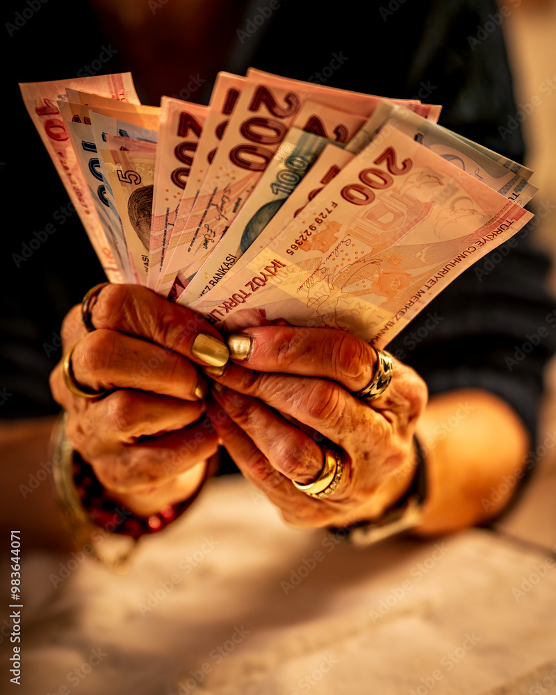 An elderly woman holding several Turkish lira banknotes, including 200 and 100 lira bills. Their hands are adorned with bracelets and rings, and they appear to be counting or organizing the money.