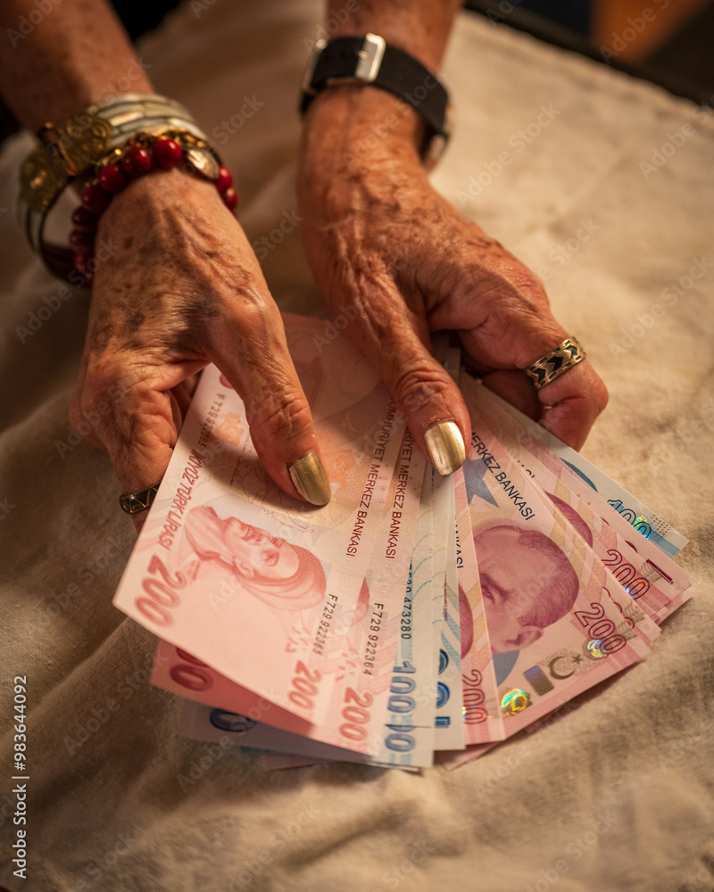 An elderly woman holding several Turkish lira banknotes, including 200 and 100 lira bills. Their hands are adorned with bracelets and rings, and they appear to be counting or organizing the money.