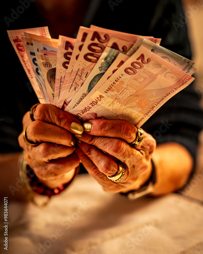An elderly woman holding several Turkish lira banknotes, including 200 and 100 lira bills. Their hands are adorned with bracelets and rings, and they appear to be counting or organizing the money.