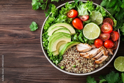 Colorful healthy salad bowl with quinoa, tomatoes, chicken, avocado, lime, and mixed greens on a wooden background from above