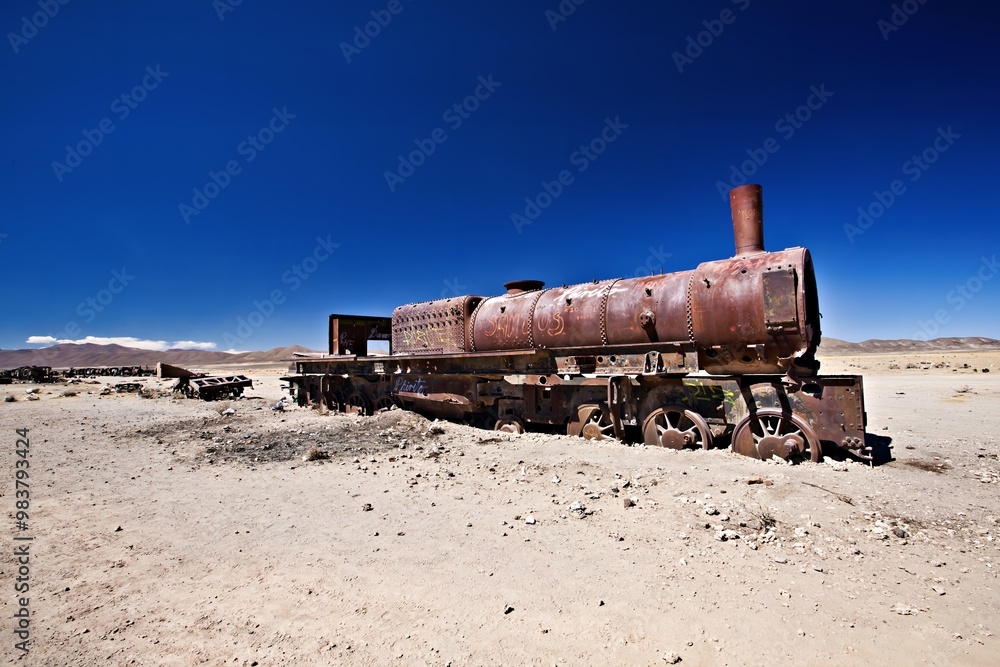 Naklejka premium An abandoned rusted steam locomotive in a desolate landscape under a clear blue sky.