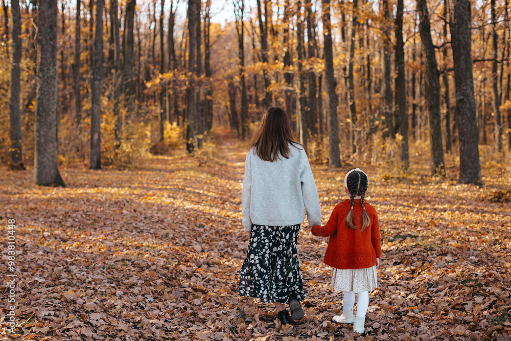 Mother and her daughter are walking in an autumn forest. The woman, dressed in a gray sweater and skirt, is walking with her daughter, who is wearing a red sweater, in the autumn forest. Back view. 
