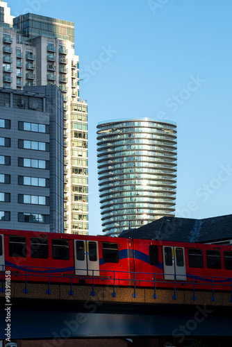 A bright red and blue train passes by towering skyscrapers in an urban setting during daylight, showcasing transportation, modernity, and city life. photo