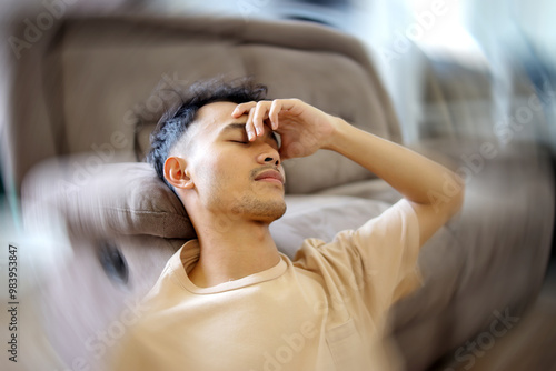 Asian Man Suddenly Feels Dizzy and Takes a Seat on Sofa. Young Guy Feeling Pain and Spinning Sensation in His Head.  photo