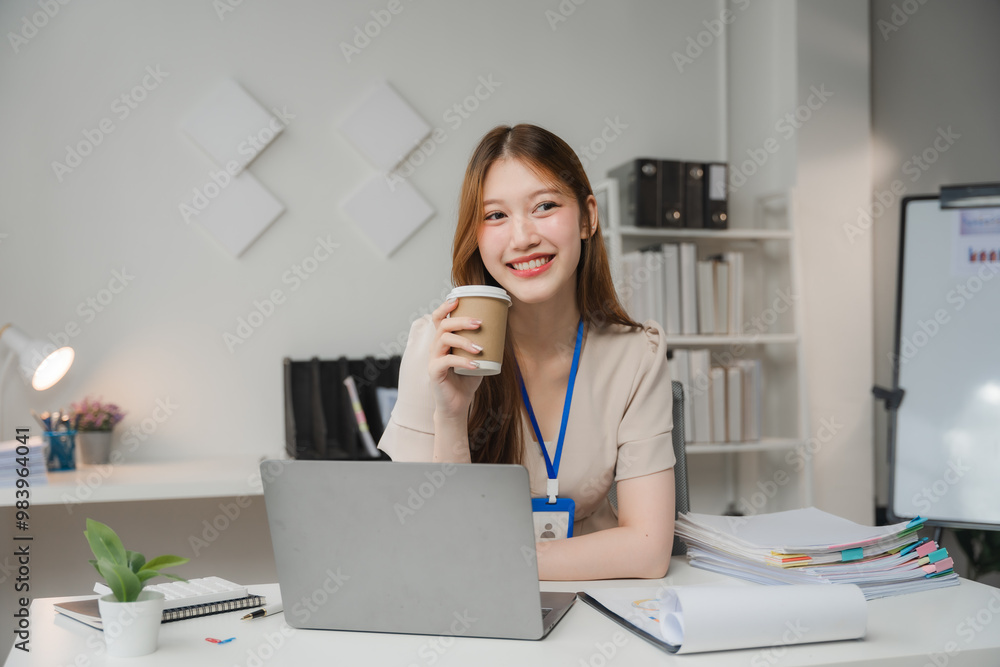 Energized Efficiency: Smiling Asian businesswoman enjoys a coffee break at her tidy office desk, embodying productivity and work-life balance Energized Efficiency: Smiling Asian businesswoman enjoys a coffee break at her tidy office desk, embodying productivity and work-life balance