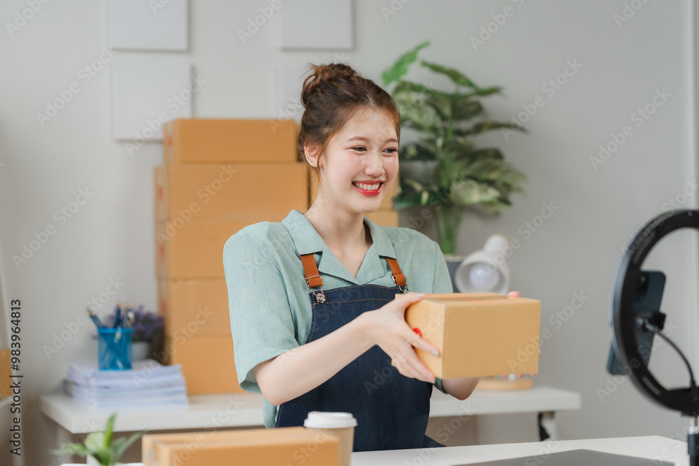 Smiling Female Entrepreneur in a Home Office Setting: A young woman in a casual green shirt and denim overalls beams with joy as she displays a cardboard box, showcasing her online business and the po Smiling Female Entrepreneur in a Home Office Setting: A young woman in a casual green shirt and denim overalls beams with joy as she displays a cardboard box, showcasing her online business and the po