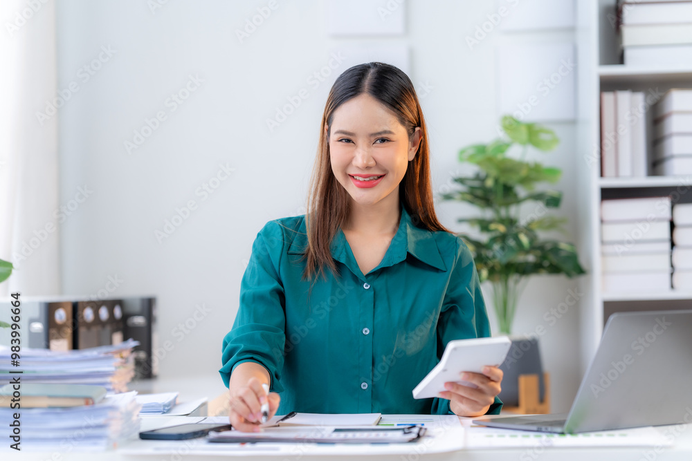 Confident Businesswoman: A professional Asian woman in a green blouse confidently works at her desk, holding a calculator and a pen. She smiles directly at the camera, showcasing her determination and Confident Businesswoman: A professional Asian woman in a green blouse confidently works at her desk, holding a calculator and a pen. She smiles directly at the camera, showcasing her determination and