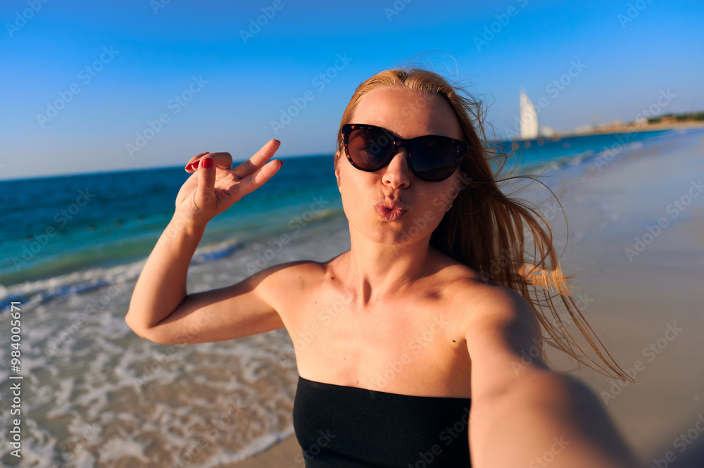 Young woman taking selfies against the sea folding her lips in a kiss and showing a peace sign. Woman taking selfie near sea under sky. Dubai, UAE, Burj Al Arab Jumeirah. Young woman taking selfies against the sea folding her lips in a kiss and showing a peace sign. Woman taking selfie near sea under sky. Dubai, UAE, Burj Al Arab Jumeirah.