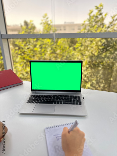 a woman watching video tutorials, making notes with a pen in an office in front of a widow with a view of tall green trees on a bright sunny day
