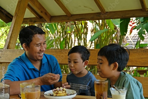 A joyful family moment of a father feeding his two sons pancakes at an outdoor setting. Perfect for content related to family bonding, parenting, breakfast promotions, or lifestyle ads emphasizing tog photo