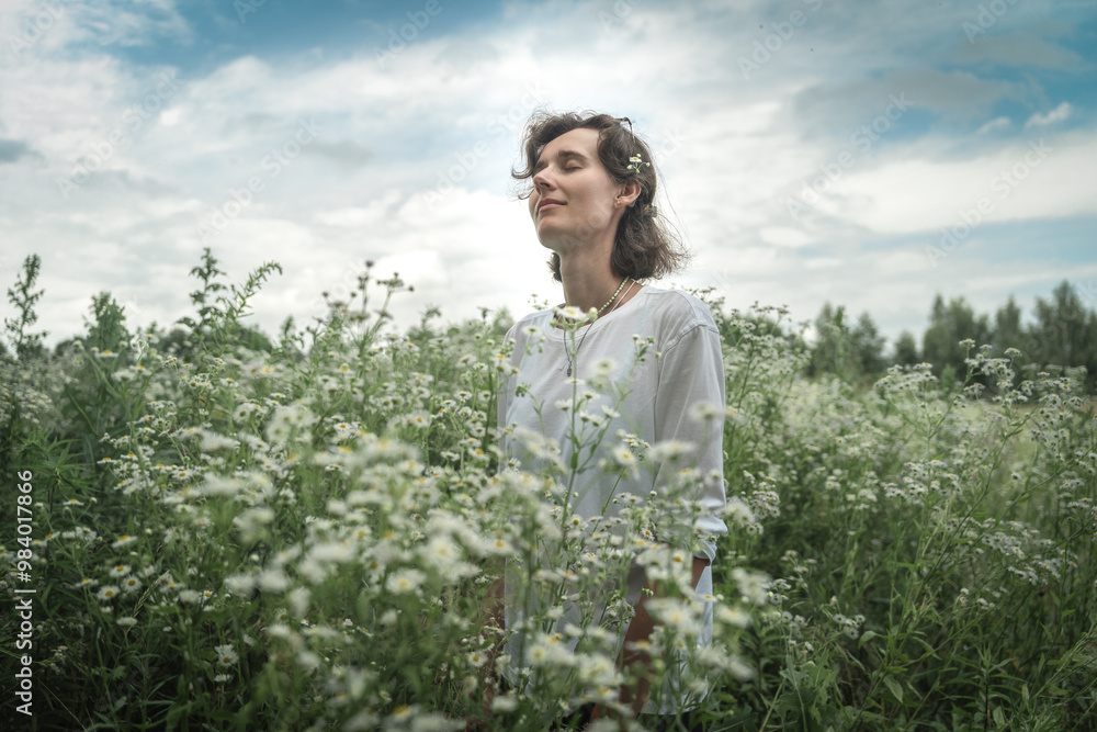 a girl of European appearance stands with her eyes closed in a chamomile field in a white T-shirt, blue sky, general plan a girl of European appearance stands with her eyes closed in a chamomile field in a white T-shirt, blue sky, general plan