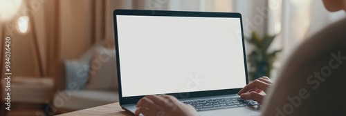 A person is typing on a laptop with a blank screen. The laptop is on a table in a living room. Mockup