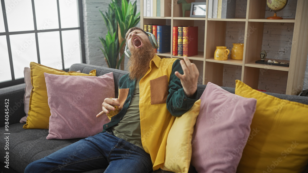 Obraz premium Bearded redhead man pretending to play guitar indoors on a sofa with colorful pillows and headphones.