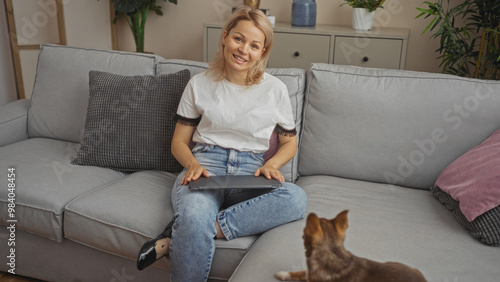 Young blonde woman sitting in the living room of her home with her laptop while her chihuahua dog looks up at her.