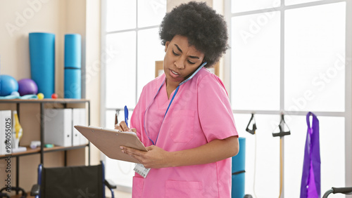 Focused african american woman in pink scrubs multitasking with clipboard and phone in rehab clinic interior. photo