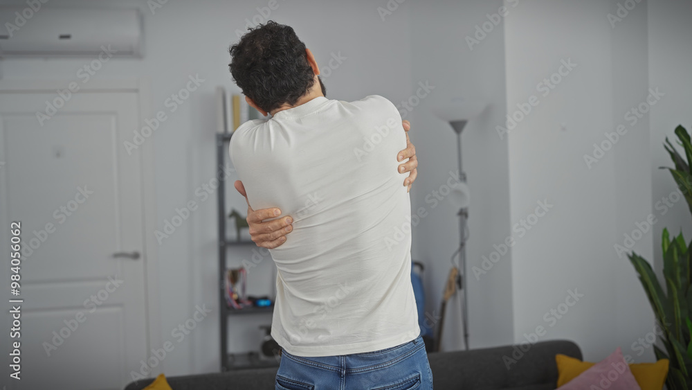 Back view of a middle-aged man with pain embracing his shoulder in a modern living room. Back view of a middle-aged man with pain embracing his shoulder in a modern living room.
