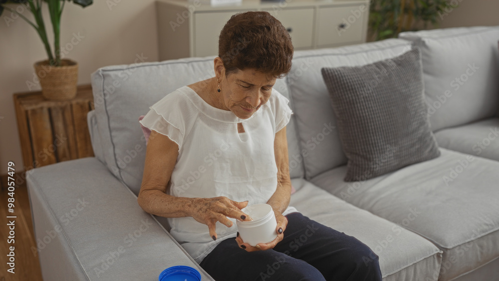 An elderly woman with short hair sits in her living room, applying cream from a jar while wearing a white blouse and sitting on a light gray sofa. An elderly woman with short hair sits in her living room, applying cream from a jar while wearing a white blouse and sitting on a light gray sofa.
