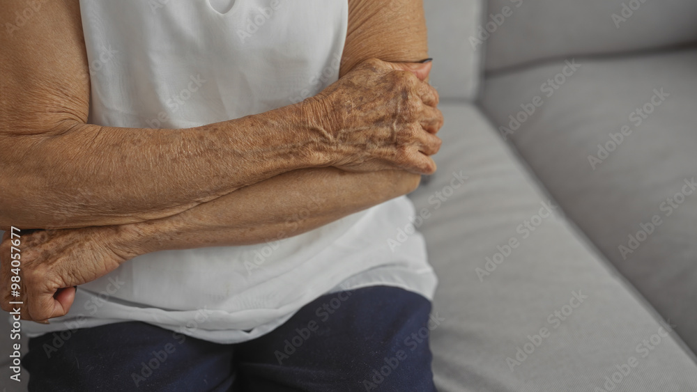 Naklejka premium An elderly woman with crossed hands sitting in a living room, highlighting the mature wrinkles and textures of her skin.