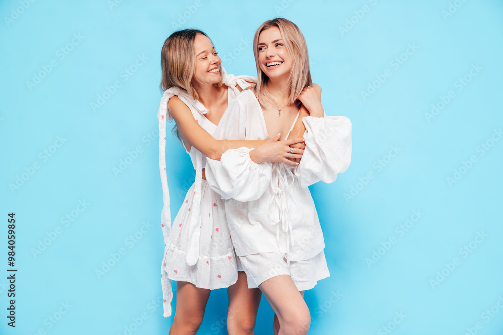 Two young beautiful smiling blond hipster female in trendy summer clothes. Blonde carefree women posing near blue wall in studio. Positive models having fun. Cheerful and happy Two young beautiful smiling blond hipster female in trendy summer clothes. Blonde carefree women posing near blue wall in studio. Positive models having fun. Cheerful and happy