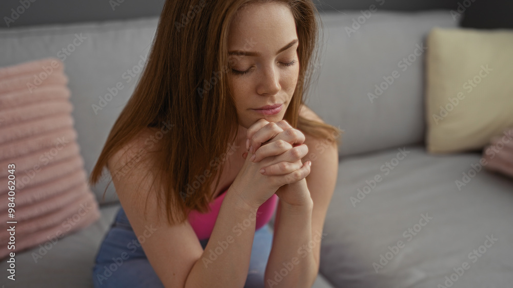Young redhead woman praying indoors with eyes closed in a cozy living room setting, conveying serenity and reflection Young redhead woman praying indoors with eyes closed in a cozy living room setting, conveying serenity and reflection