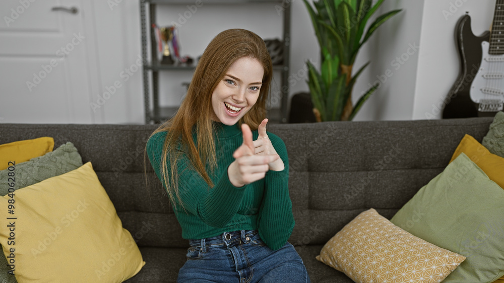 A smiling caucasian woman with blonde hair at home sitting on a sofa pointing at camera