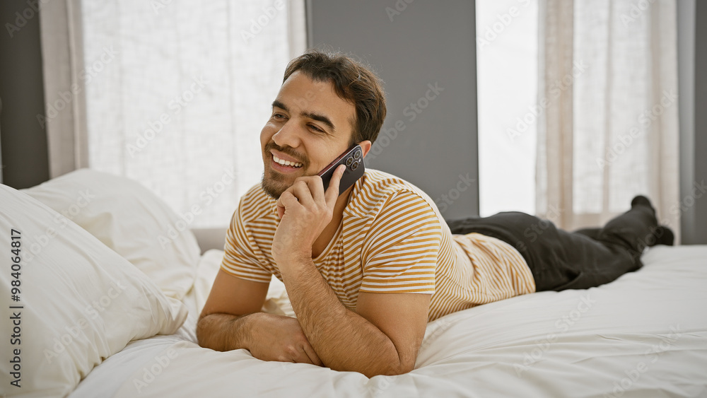 A smiling young hispanic man with a beard lying on a white bed while talking on the phone in a bedroom interior. A smiling young hispanic man with a beard lying on a white bed while talking on the phone in a bedroom interior.