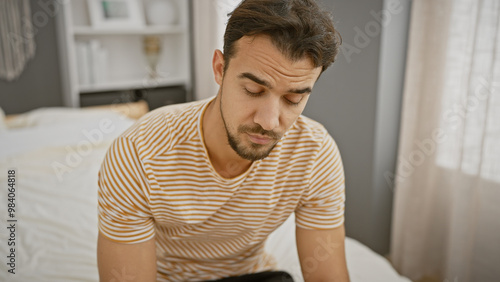 Young hispanic man with beard in striped shirt sitting pensively on bed in a bedroom interior.