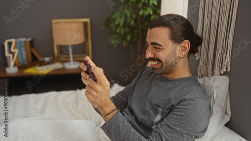 Young hispanic man with moustache relaxing in bed, smiling while using a smartphone in a cozy bedroom with decorative items and indoor plants in the background