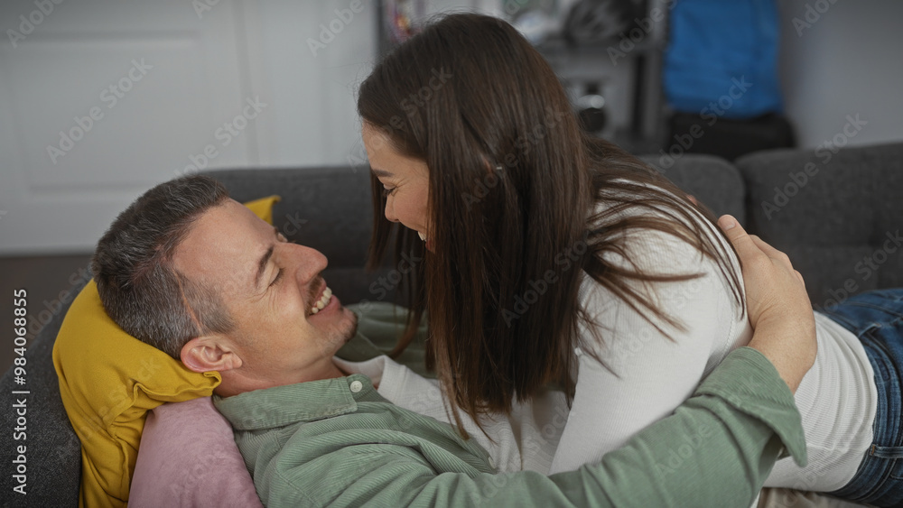 A loving couple smiles and embraces on a gray sofa, depicting affection and happiness in a cozy home setting. A loving couple smiles and embraces on a gray sofa, depicting affection and happiness in a cozy home setting.