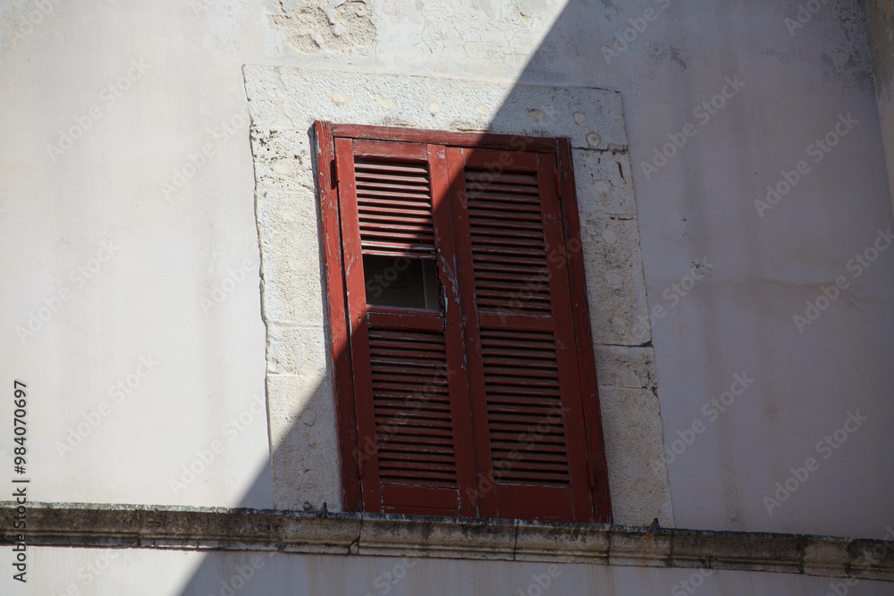 Weathered red wooden window shutters on a white stucco wall, partially open, casting dramatic shadows in bright sunlight. Urban architectural detail with rustic Mediterranean charm.
Weathered red wooden window shutters on a white stucco wall, partially open, casting dramatic shadows in bright sunlight. Urban architectural detail with rustic Mediterranean charm.
