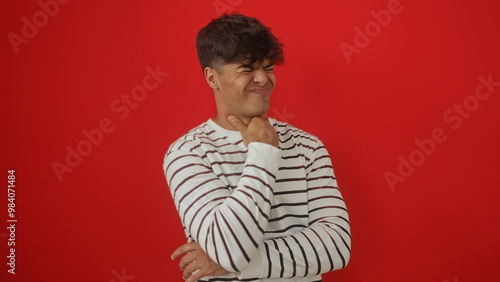 Hispanic young man in a striped sweater feeling throat pain while standing against a vibrant red background.