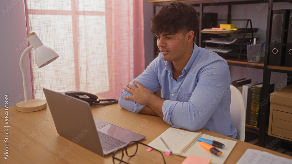 Young hispanic man in a blue shirt working at a desk in an office with a laptop, notebook, and desk lamp in a well-lit room. Young hispanic man in a blue shirt working at a desk in an office with a laptop, notebook, and desk lamp in a well-lit room.