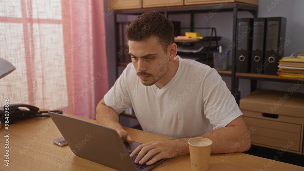 A handsome young hispanic man with a beard works intently on a laptop in a cluttered office, highlighted by bright natural sunlight and various office supplies.