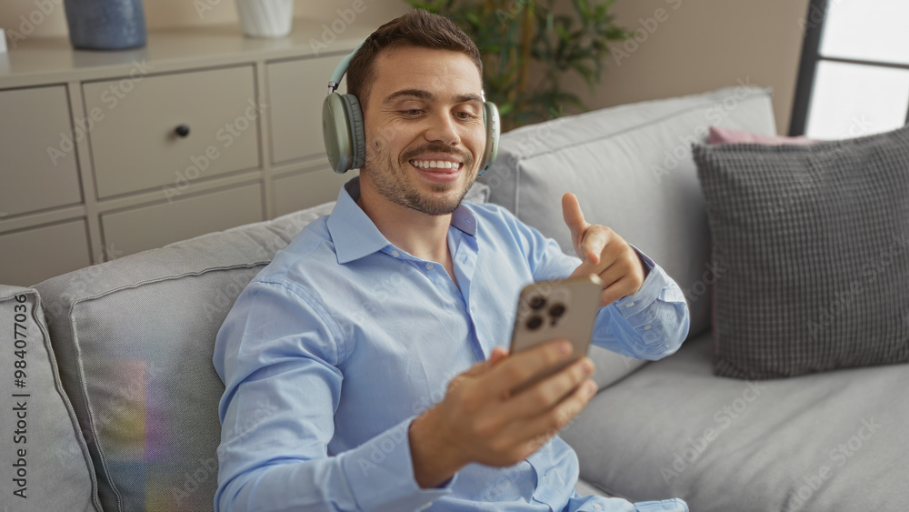 A young hispanic man with a beard is smiling and wearing headphones while using his smartphone in a modern living room at home. A young hispanic man with a beard is smiling and wearing headphones while using his smartphone in a modern living room at home.